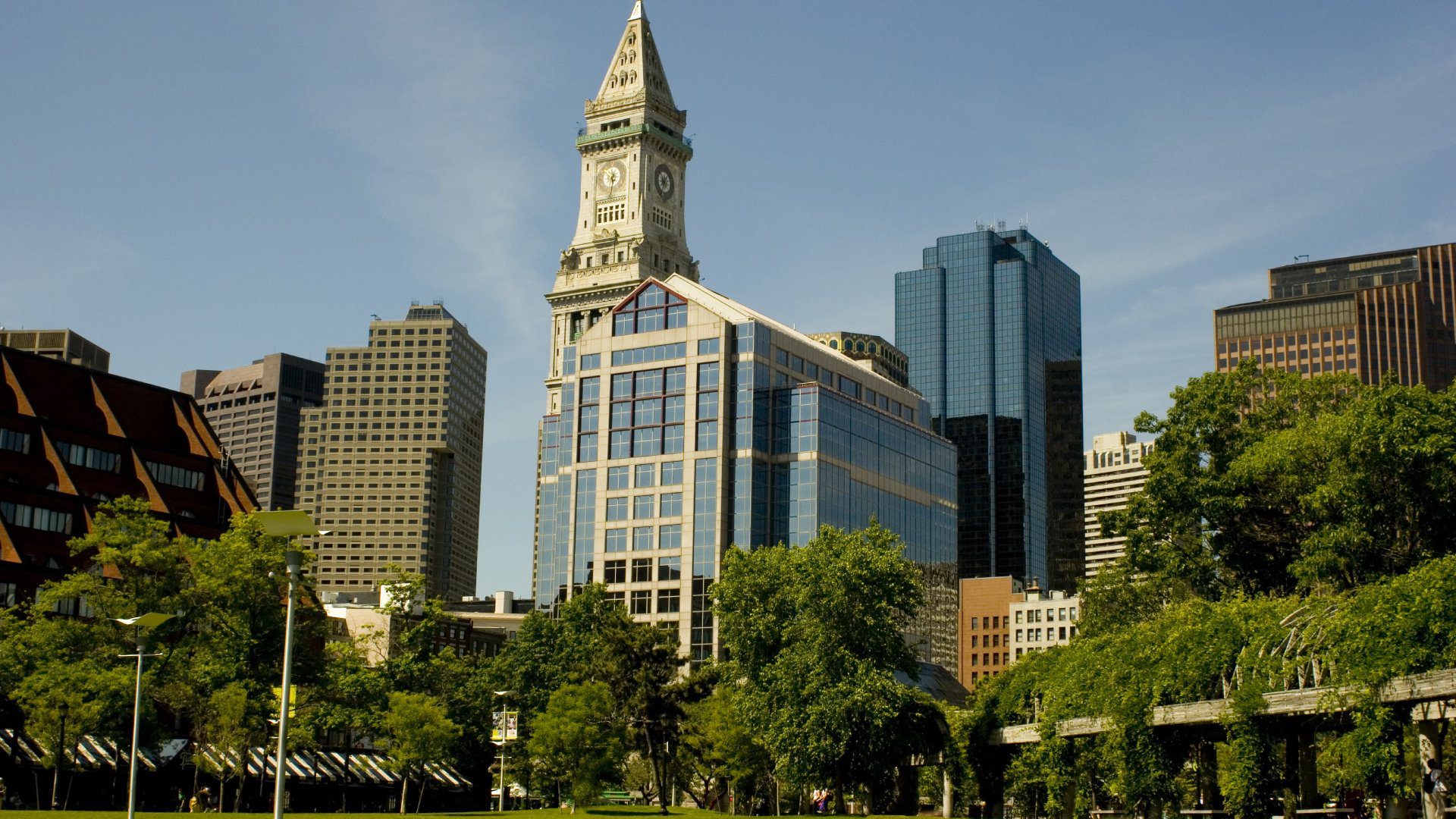Cityscape with a mix of modern and historical buildings, dominated by a clock tower in the center. Green trees and a park area are in the foreground under a clear sky.