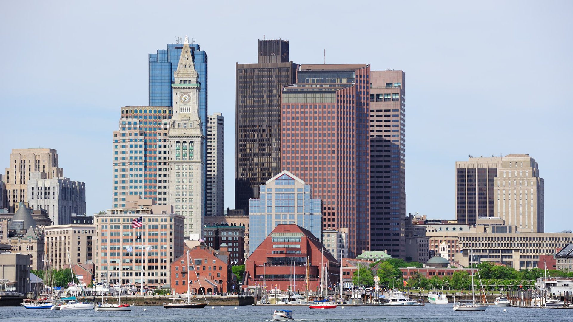 City skyline with modern and historic buildings by the waterfront, featuring boats on the water and a clear sky.