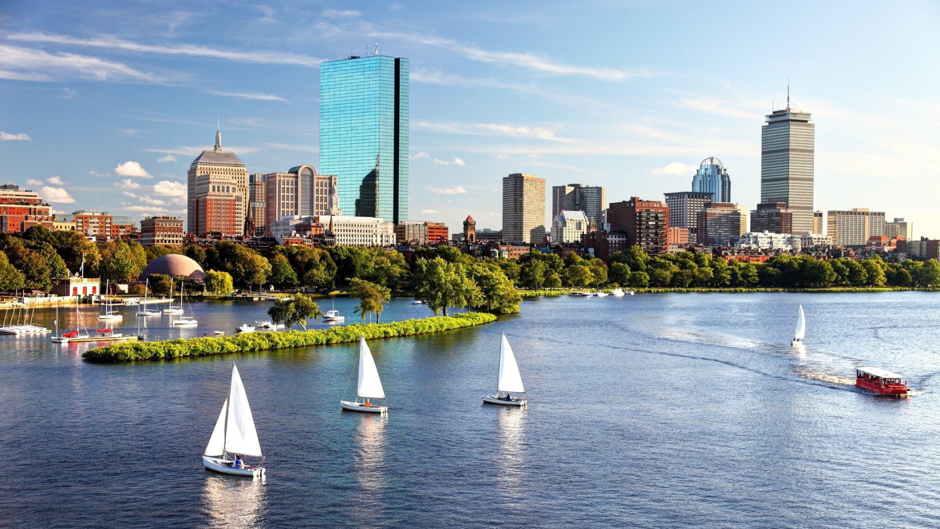 Skyline of Boston with sailboats on the Charles River. Prominent buildings and greenery are visible under a clear blue sky.
