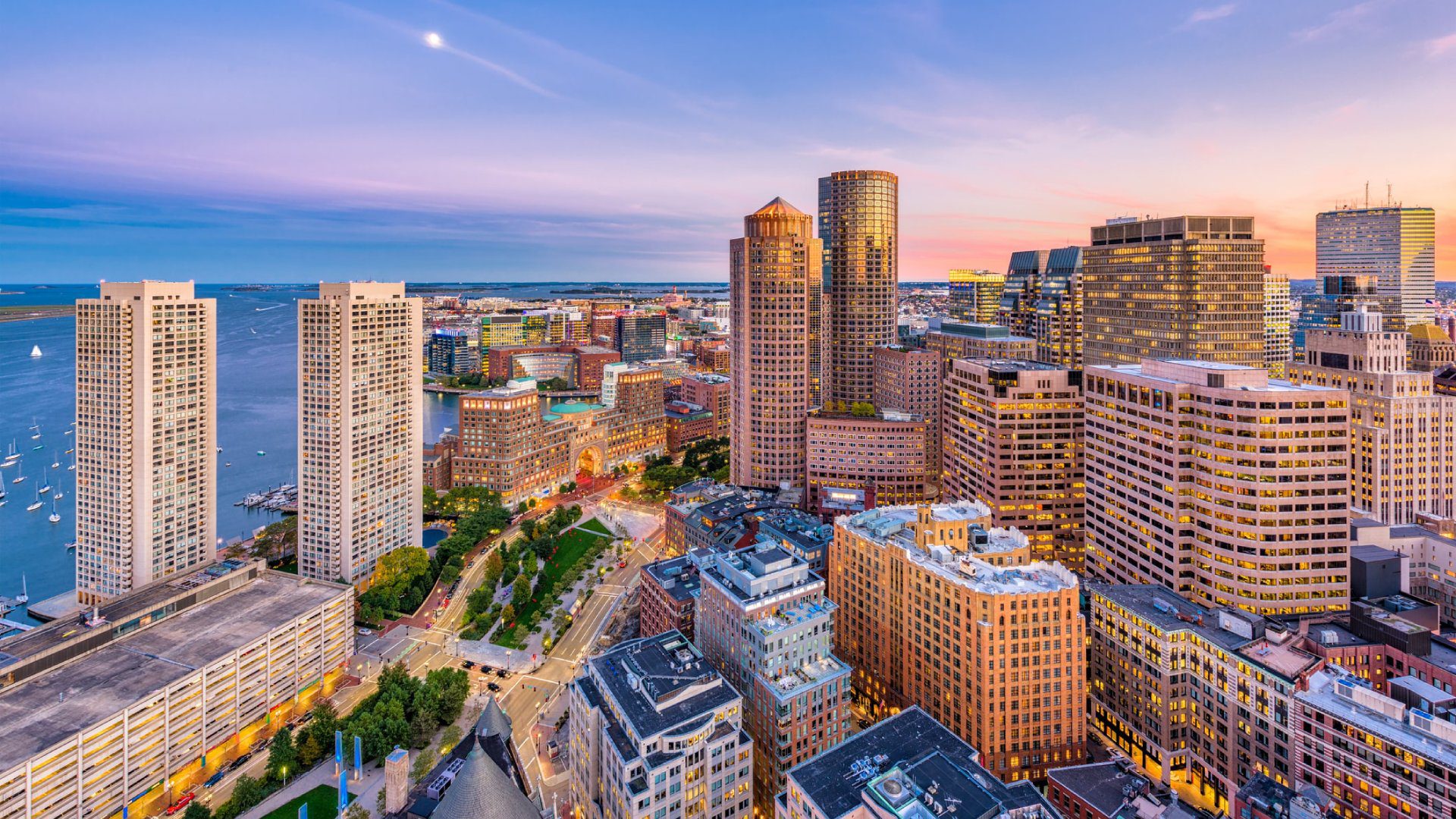 Aerial view of a cityscape at sunset featuring skyscrapers and buildings with water and boats in the background under a colorful sky.