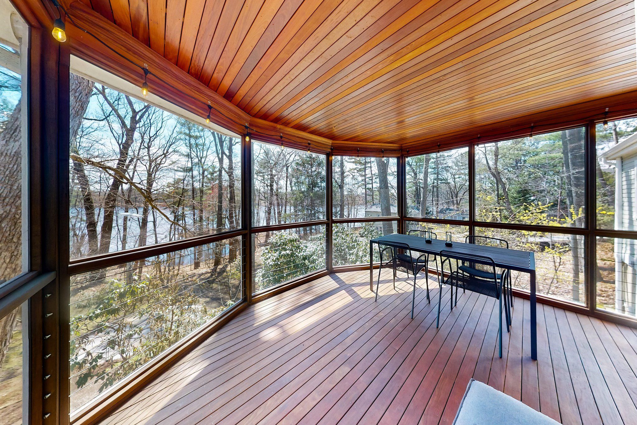 A cozy screened porch with wooden flooring and ceiling overlooking a wooded area.