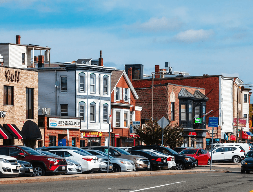 A busy street with parked cars and small businesses under a clear sky.
